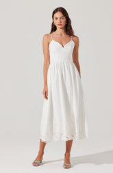 A woman is wearing a white, sleeveless, ankle-length dress with thin spaghetti straps. The dress has a fitted bodice with a V-shaped neckline and a gathered waist that flows into a full skirt. The skirt features eyelet embroidery near the hem, adding a delicate decorative detail. She is also wearing open-toed, strappy high-heeled sandals in a light color.